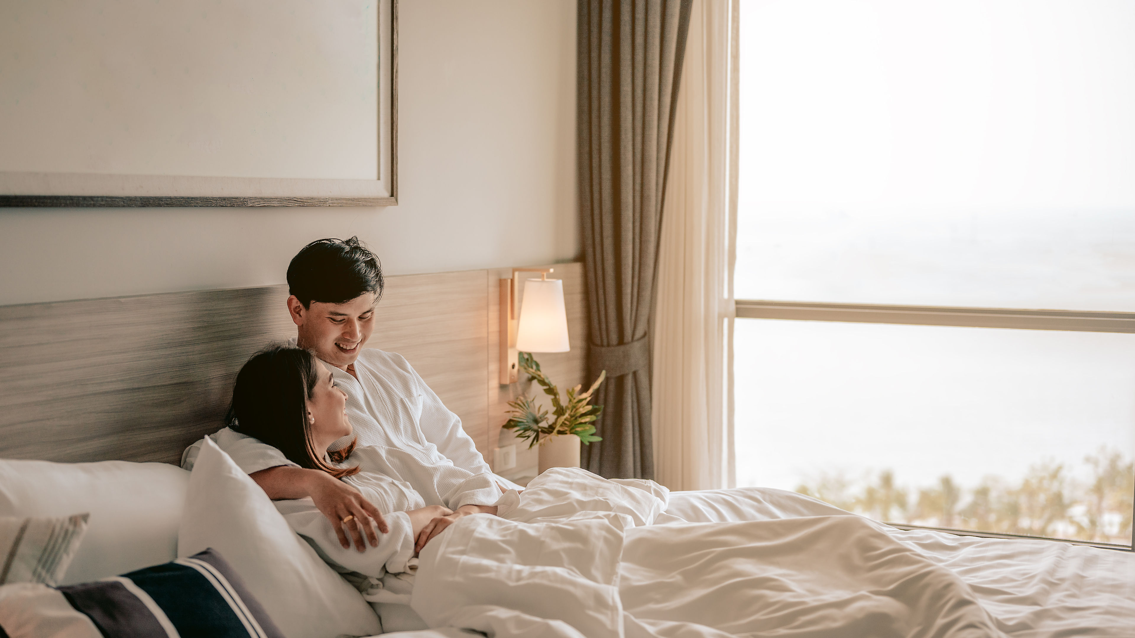 Asian couple is hugging and smiling sitting on the bed in a bedroom evening.